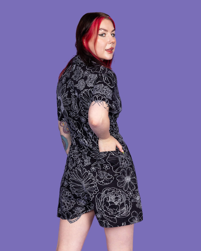 Close up of the Black Floral Stretch Twill Playsuit worn by a femme model with mid length red and black hair on a purple studio background. They are facing away with one hand resting in the back pocket looking over their right shoulder smiling to camera. The black base playsuit has white floral outlines all over.