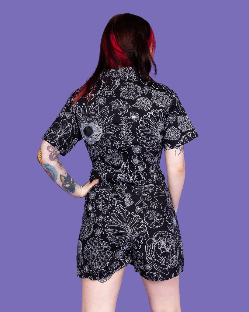 Close up of the Black Floral Stretch Twill Playsuit worn by a femme model with mid length red and black hair on a purple studio background. They are facing away with one hand on their hip. The black base playsuit has white floral outlines all over.