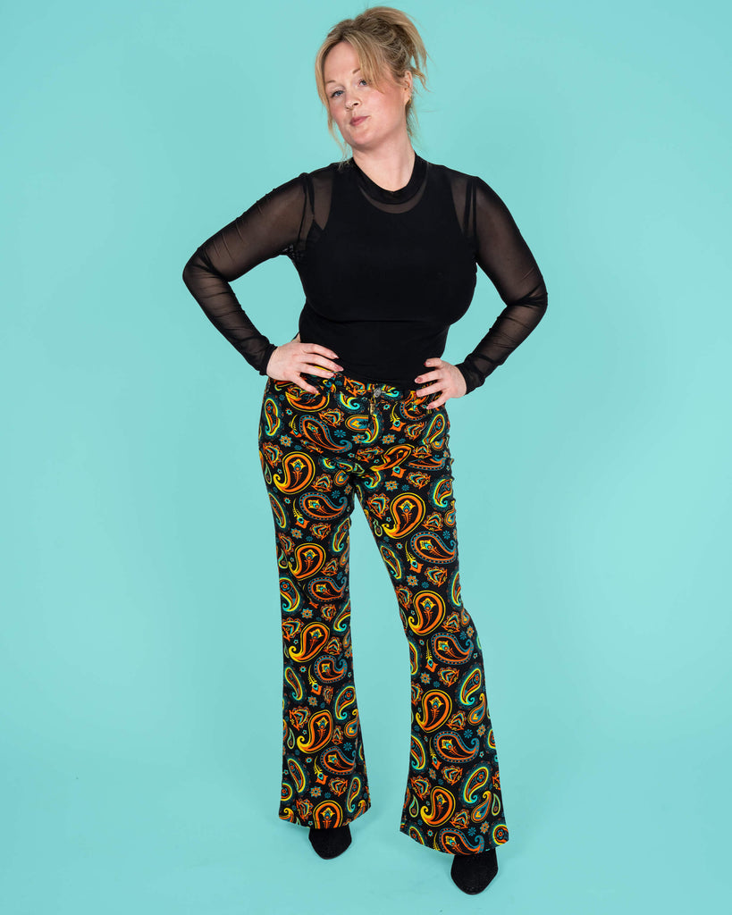 The model confidently poses in Run & Fly's black paisley flares, showcasing the high-waisted retro design. The bold orange, turquoise, and yellow paisley print exudes vintage charm while supporting ethical slow fashion.