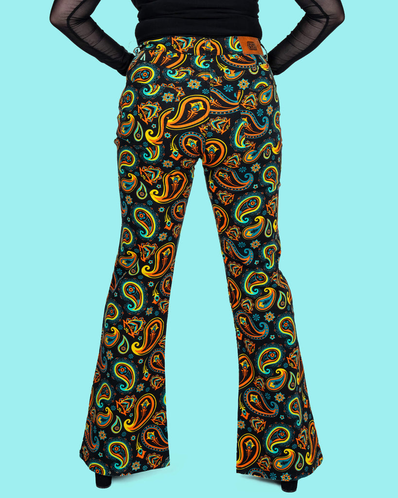 Half length back view of Run & Fly's black paisley flares, styled with a black mesh top. The bold retro-inspired paisley print in orange, turquoise, and yellow tones stands out, making these ethically made trousers a vibrant addition to any wardrobe.