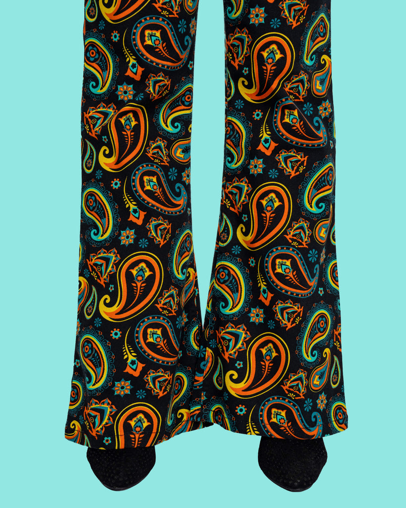 A detailed view of the vibrant paisley print at the hem of the black flared trousers by Run & Fly. Featuring retro-style orange, turquoise, and yellow paisley patterns with floral accents, these ethically made flares add a bold vintage vibe.