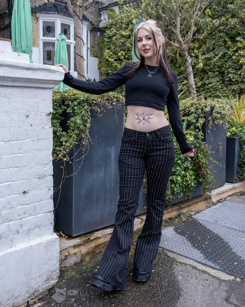 Model wearing the Run & Fly Black & White Pinstripe Bell Bottom Stretch Flares in a garden-style urban setting, showing off the flattering high waist and leg-lengthening flare.