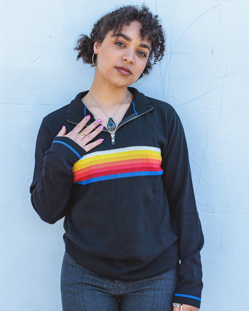 Georgia a black model with curly hair is smiling wearing a retro knit high neck Run & Fly jumper with striped detail on the front. She is stood outside leaning against a white wall.