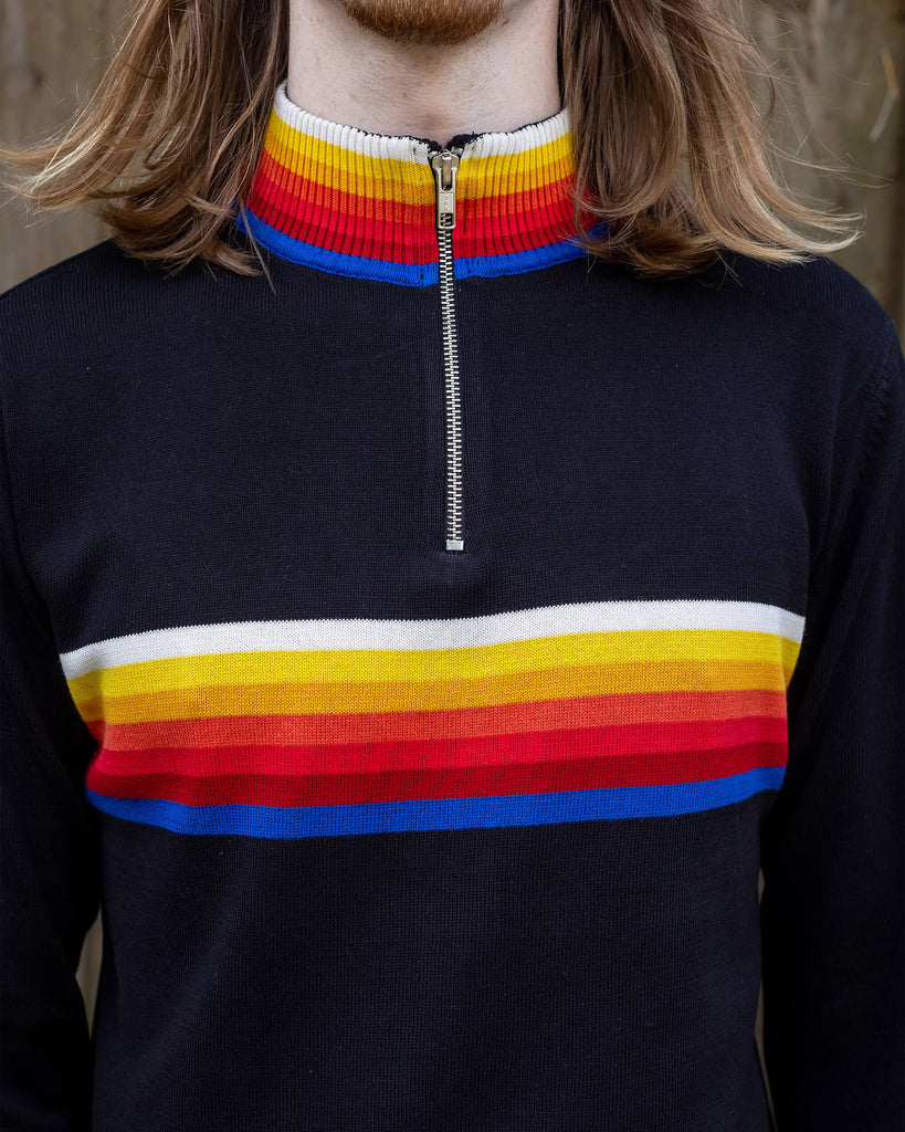 Close up of Jack, a man in his 20's with shoulder length hair and a little beard wearing glasses and a retro knit high neck Run & Fly jumper with striped detail on the front. He is stood outside against a fence.
