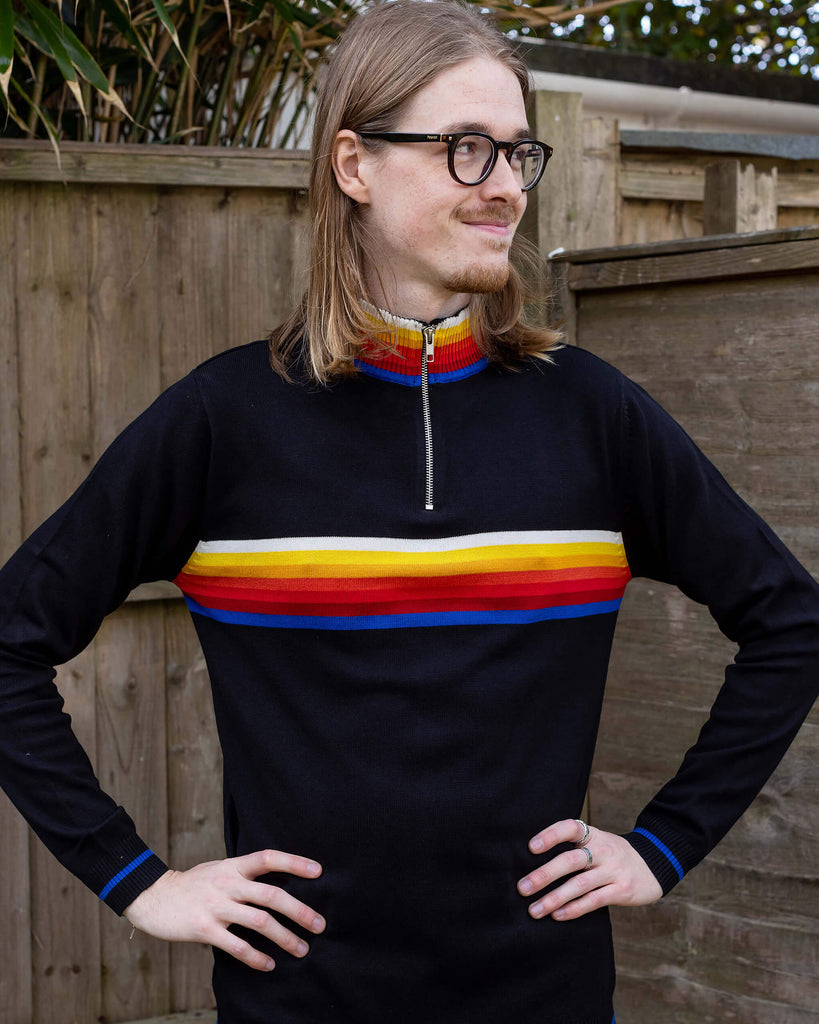 Jack is a man in his 20's with shoulder length hair and a little beard wearing glasses and a retro knit high neck Run & Fly jumper with striped detail on the front. He is stood outside against a fence smiling looking right with his hands on his hips.