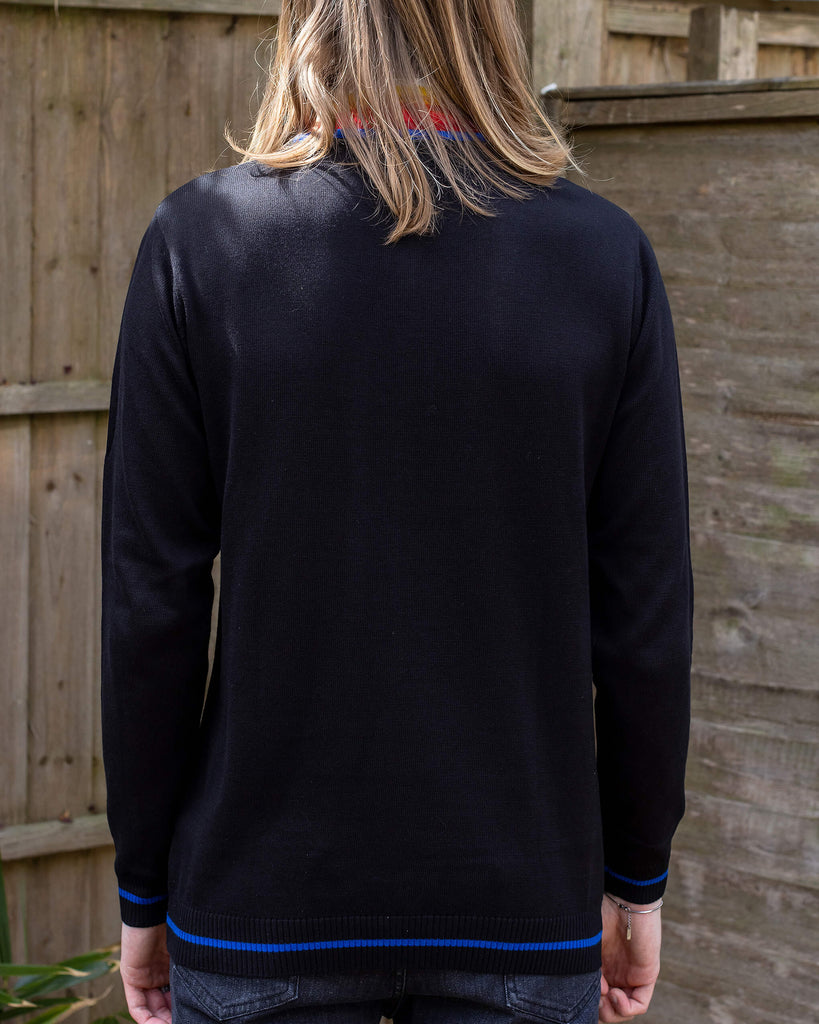 Jack is a man in his 20's with shoulder length hair and a little beard wearing glasses and a retro knit high neck Run & Fly jumper with striped detail on the front. He is stood outside against a fence facing away to show the plain back.