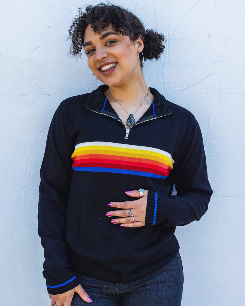 Georgia a black model with curly hair is smiling wearing a retro knit high neck Run & Fly jumper with striped detail on the front. She is stood outside leaning against a white wall smiling.