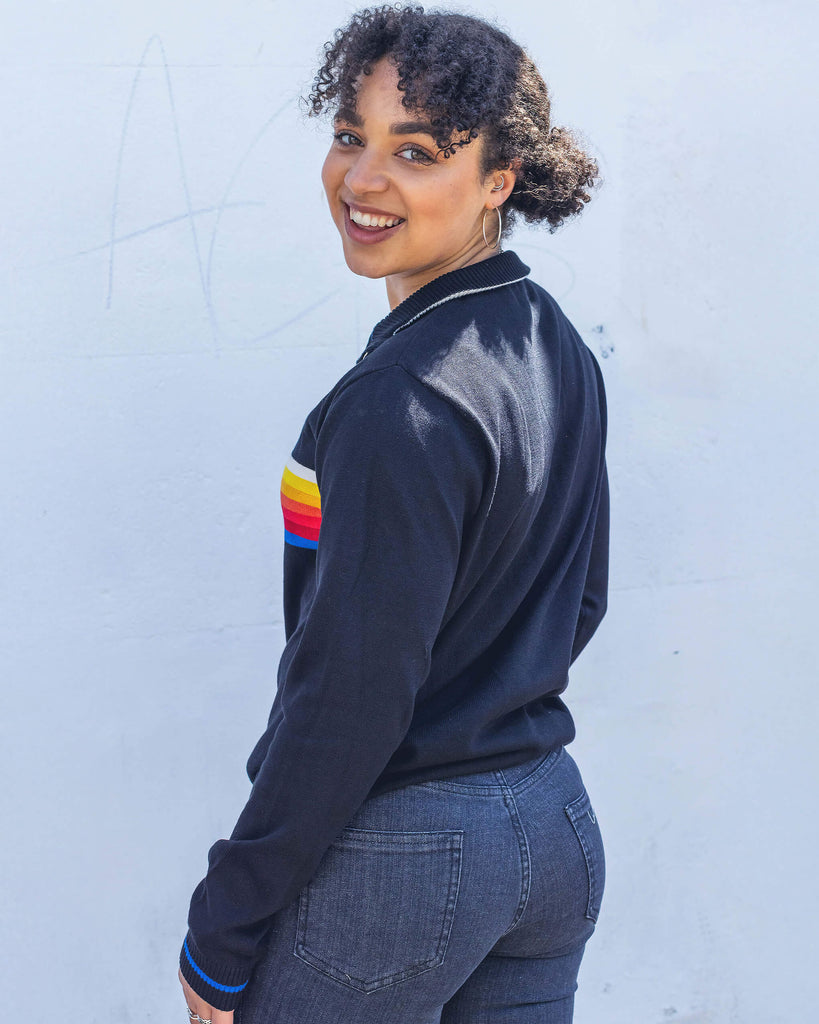 Georgia a black model with curly hair is smiling wearing a retro knit high neck Run & Fly jumper with striped detail on the front. She is stood outside leaning against a white wall smiling.