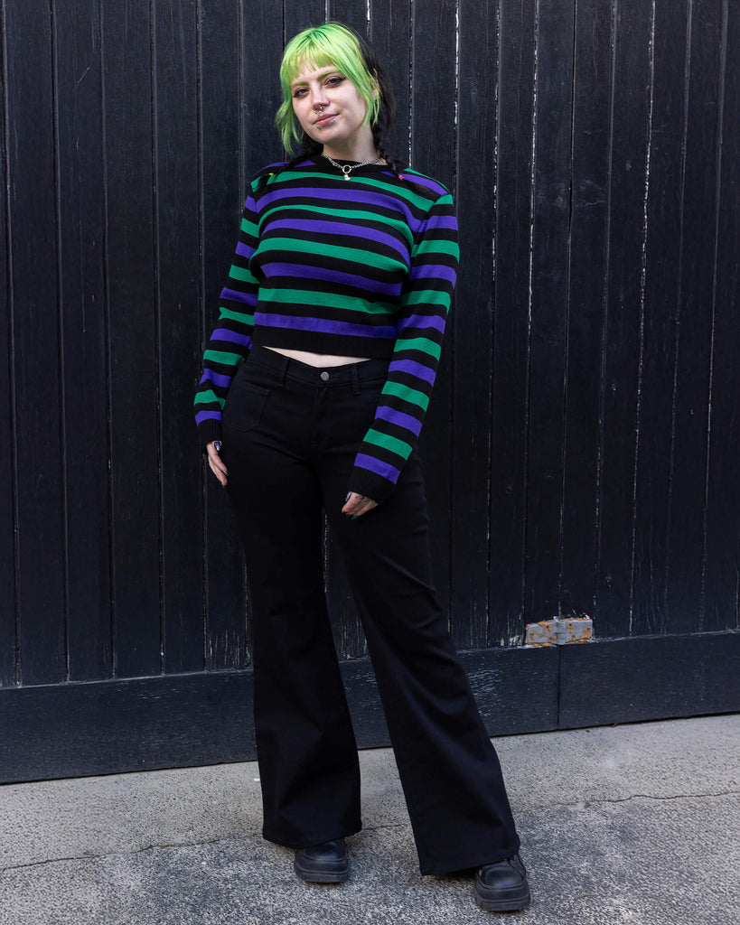 Model with green and black hair wearing Black Striped Purple and Green Cropped Jumper paired with black flared. jeans and chunky black boots. The jumper is a cropped style and has a black background colour with chunky muted green and purple horizontal stripes across it. 