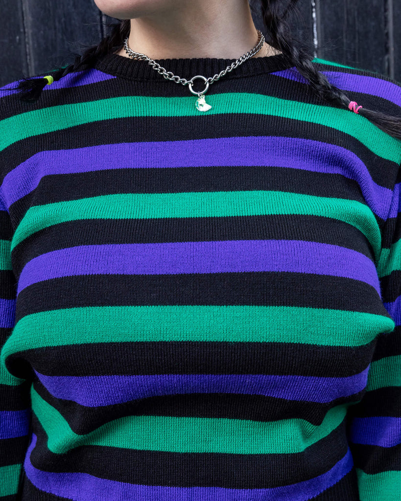 Model with green and black hair wearing Black Striped Purple and Green Cropped Jumper paired with black jeans. The jumper is a cropped style and has a black background colour with chunky muted green and purple horizontal stripes across it. 