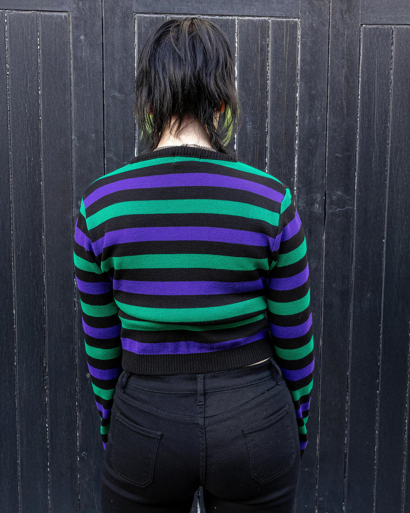Model with green and black hair wearing Black Striped Purple and Green Cropped Jumper paired with black jeans. The jumper is a cropped style and has a black background colour with chunky muted green and purple horizontal stripes across it. 
