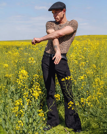 Model is stood in a yellow flower field wearing black corduroy bell bottom super flares with a natural leopard print t-shirt, black beret cap and black trainers. They are facing the camera and looking off to the left with their arms crossed over.