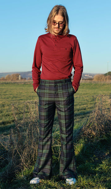 Jack is stood in front of a grassy field wearing the blackwatch tartan plaid bell bottom trousers with a long sleeve red tshirt and white trainers. They are facing the camera and looking down with both hands in their pockets.