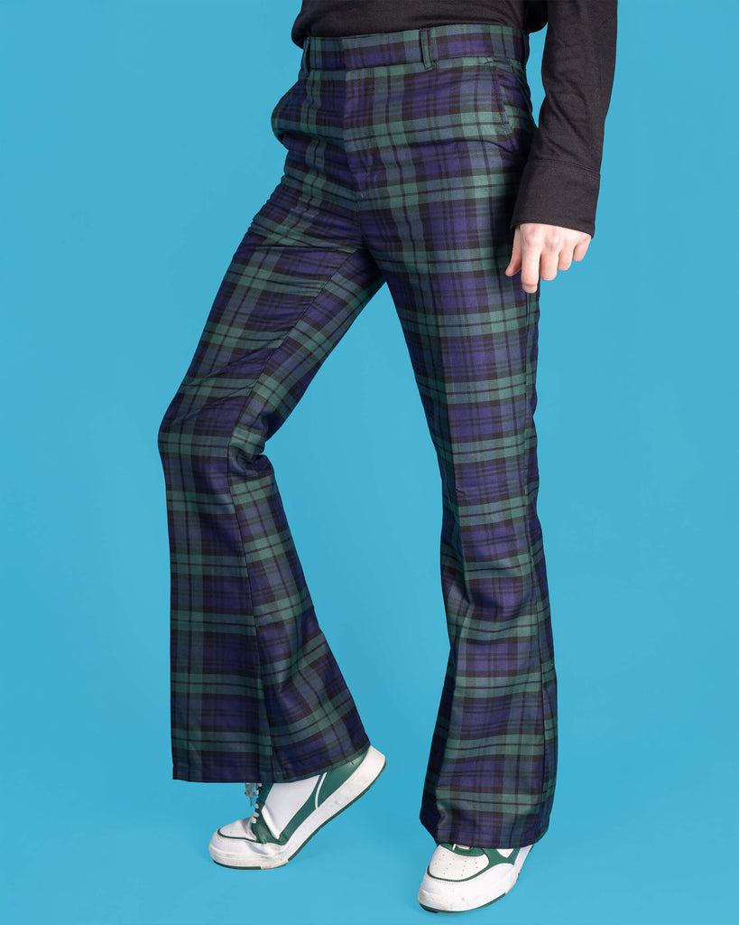 Jack is stood in front of a blue studio background wearing the black watch tartan plaid bell bottom trousers with green and navy tones, paired with white trainers and a black long sleeve top. He is facing to the left with one leg bent forward to highlight the bell bottom flares. Photo is cropped from the waist down.