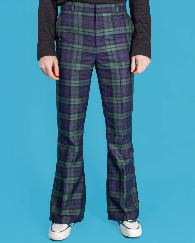 Jack is stood in front of a blue studio background wearing the black watch tartan plaid bell bottom trousers with green and navy tones, paired with white trainers and a black long sleeve top. They are facing forward with both arms resting by their side. Photo is cropped from the waist down.
