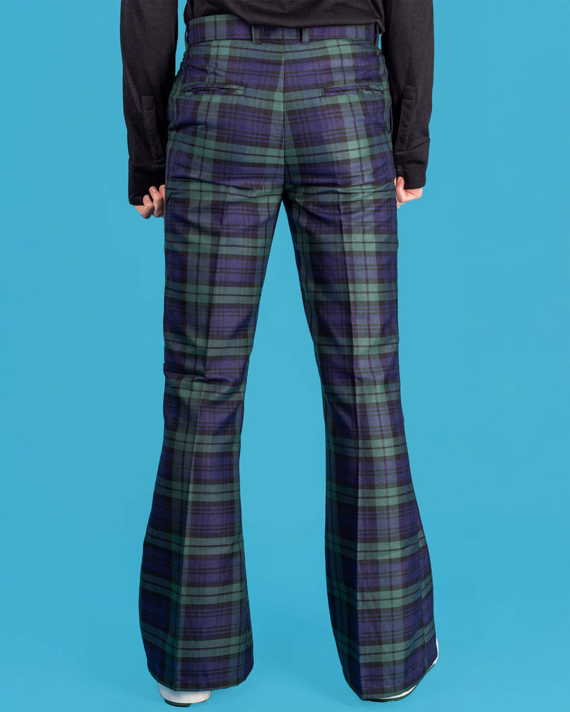 Jack is stood in front of a blue studio background wearing the black watch tartan plaid bell bottom trousers with green and navy tones, paired with white trainers and a black long sleeve top. They are facing away from the camera with both arms resting by their sides. Photo is cropped from the waist down.