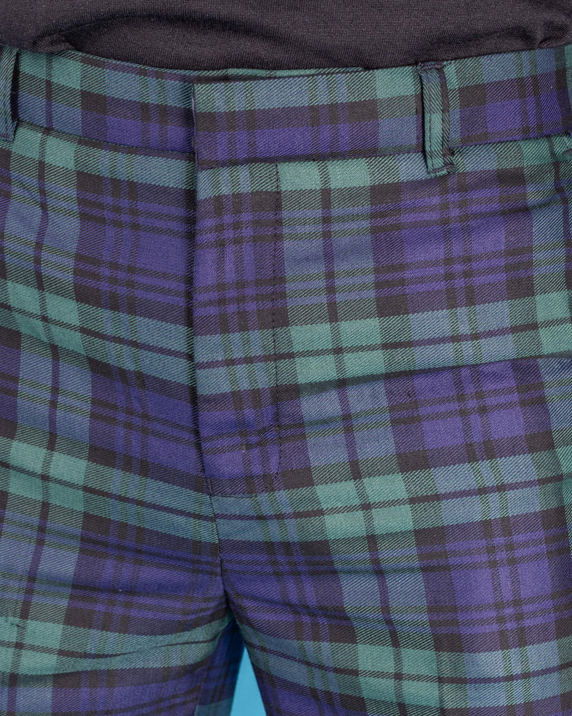 Jack is stood in front of a blue studio background wearing the black watch tartan plaid bell bottom trousers with green and navy tones paired with a black top. Photo is cropped in on the front zip fly close and belt loops.