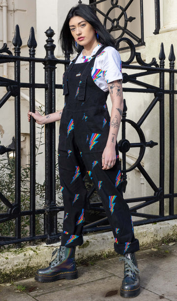 Gabriella stands in front of black railings in Brighton wearing a black lightning bolt super stretch twill dungarees, paired with a white lightning bolt tshirt and grey chunky dr martens. Model is looking into the camera with one hand on the railings