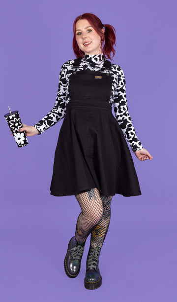 model with red hair wearing a black flared pinafore paired with a long sleeve cow print turtle neck, fishnet tights and boots. The model is smiling and holding a black and white floral travel cup in front of a purple background.
