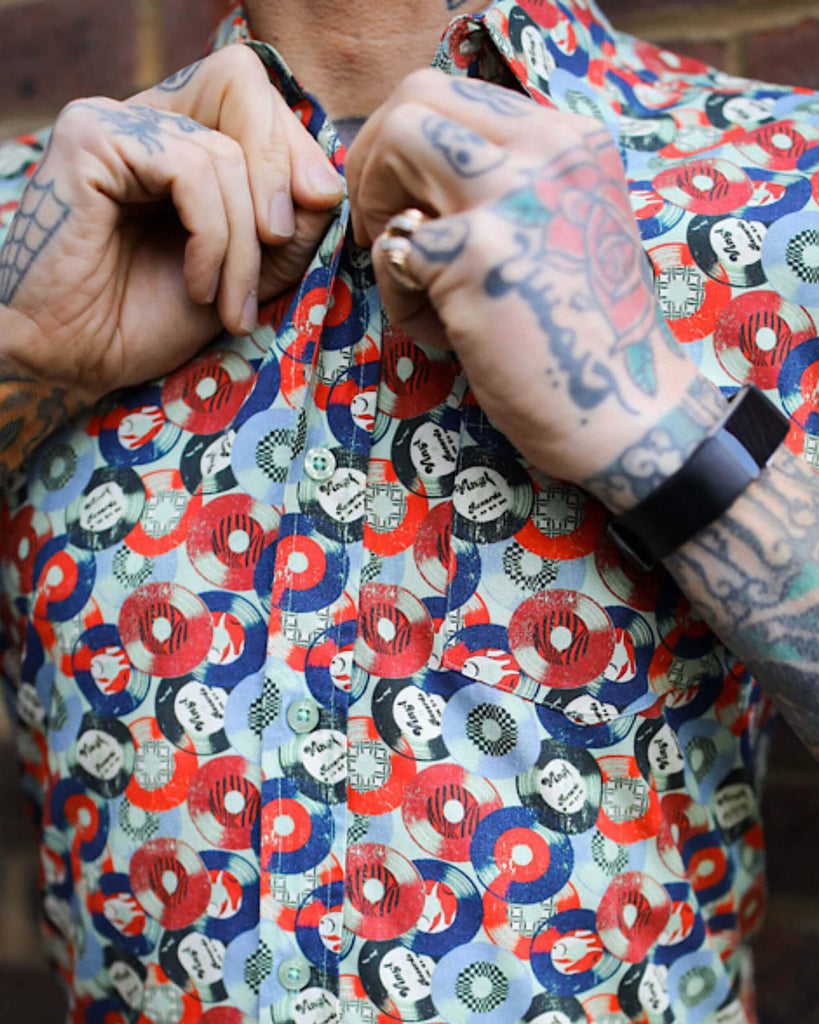 Jim is stood in front of a brick wall wearing the red and blue record short sleeve shirt with blue denim jeans. They are heavily tattooed with swept back silver hair. They are buttoning up the top button and photo is cropped in on the chest.