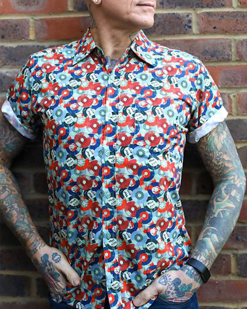 Jim is stood in front of a brick wall wearing the red and blue record short sleeve shirt with blue denim jeans. They are heavily tattooed with swept back silver hair. They are facing the camera posing with both hands in their front jean pockets whilst looking off to the right. Photo is cropped from the hips up.