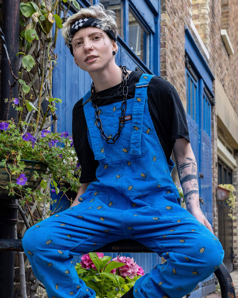 Jamie a white Masc trans man with blonde short hair is wearing blue corduroy bee printed dungarees, black boots and a black tee with big black chains. He is sat on a railing in a mews in Hove shot by Amy Davies Photography