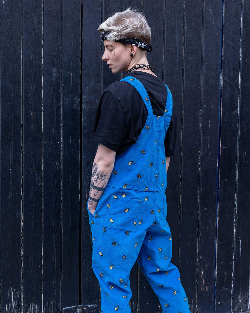 Back shot of Jamie a white Masc trans man with blonde short hair is wearing blue corduroy bee printed dungarees, black boots and a black tee with big black chains. He is leaning on a black door in a mews in Hove shot by Amy Davies Photography