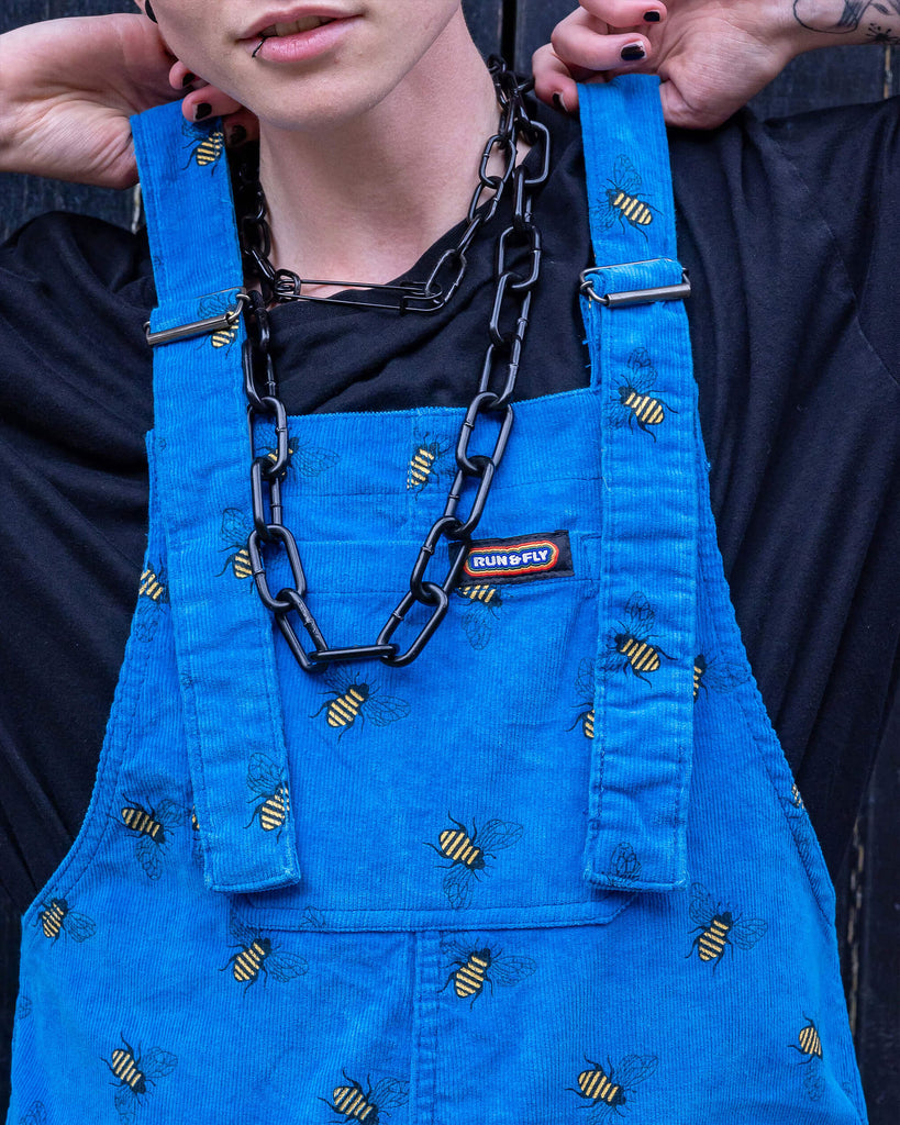 Jamie a white Masc trans man with blonde short hair is wearing blue corduroy bee printed dungarees, black boots and a black tee with big black chains. He is leaning on a black door in a mews in Hove shot by Amy Davies Photography