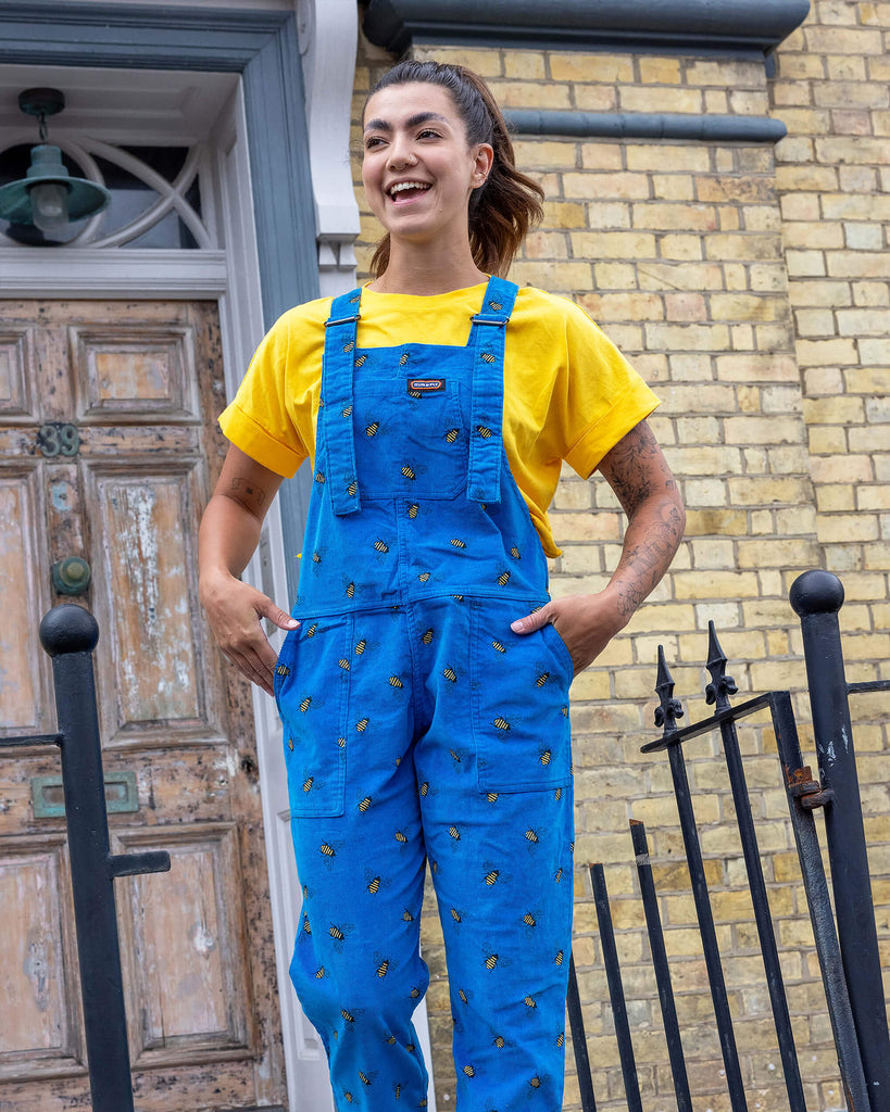 Sophie a tanned white model with hair up and tattoos on her arm is looking away from camera laughing wearing blue corduroy bee dungarees with a yellow tee and black boots shot in Hove