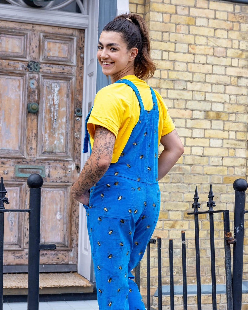 Sophie a tanned white model with hair up and tattoos on her arm is looking at the camera laughing wearing blue corduroy bee dungarees with a yellow tee and black boots shot in Hove