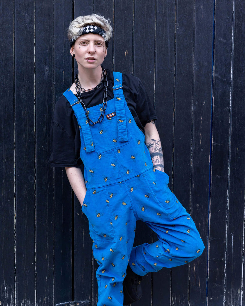 Jamie a white Masc trans man with blonde short hair is wearing blue corduroy bee printed dungarees, black boots and a black tee with big black chains. He is leaning on a black door in a mews in Hove shot by Amy Davies Photography