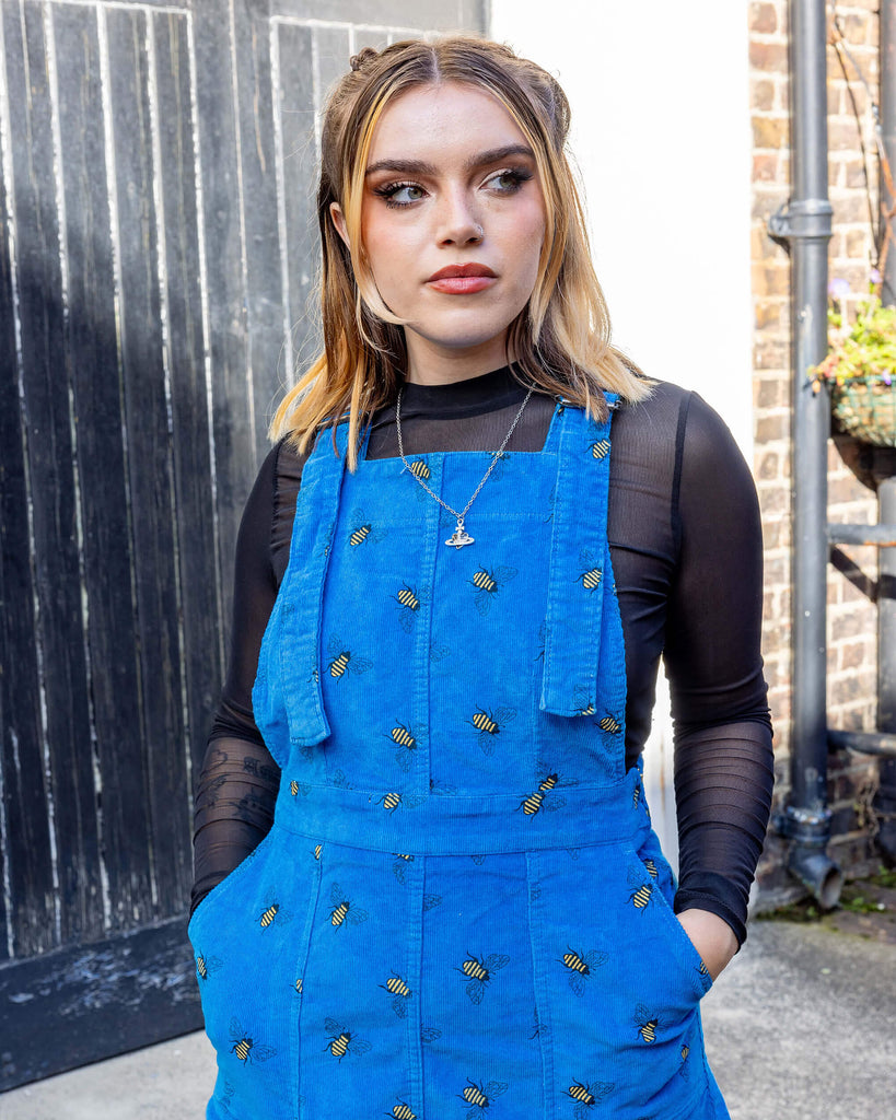 white femme model with blonde hair is wearing Run & Fly Blue Bee Stretch Corduroy Pinafore Dress paired with a long sleeve black mesh top. The bright blue pinafore has an all over bee print. The model is looking into the distance with her hands in the pockets of the pinafore outdoors in Hove.