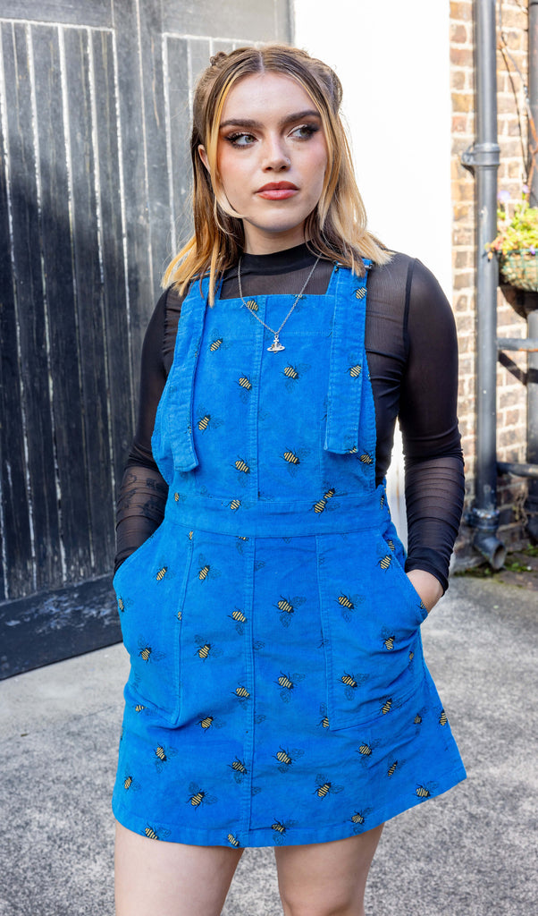 white femme model with blonde hair is wearing Run & Fly Blue Bee Stretch Corduroy Pinafore Dress paired with a long sleeve black mesh top. The bright blue pinafore has an all over bee print. The model is looking into the distance with her hands in the pockets of the pinafore outdoors in Hove.