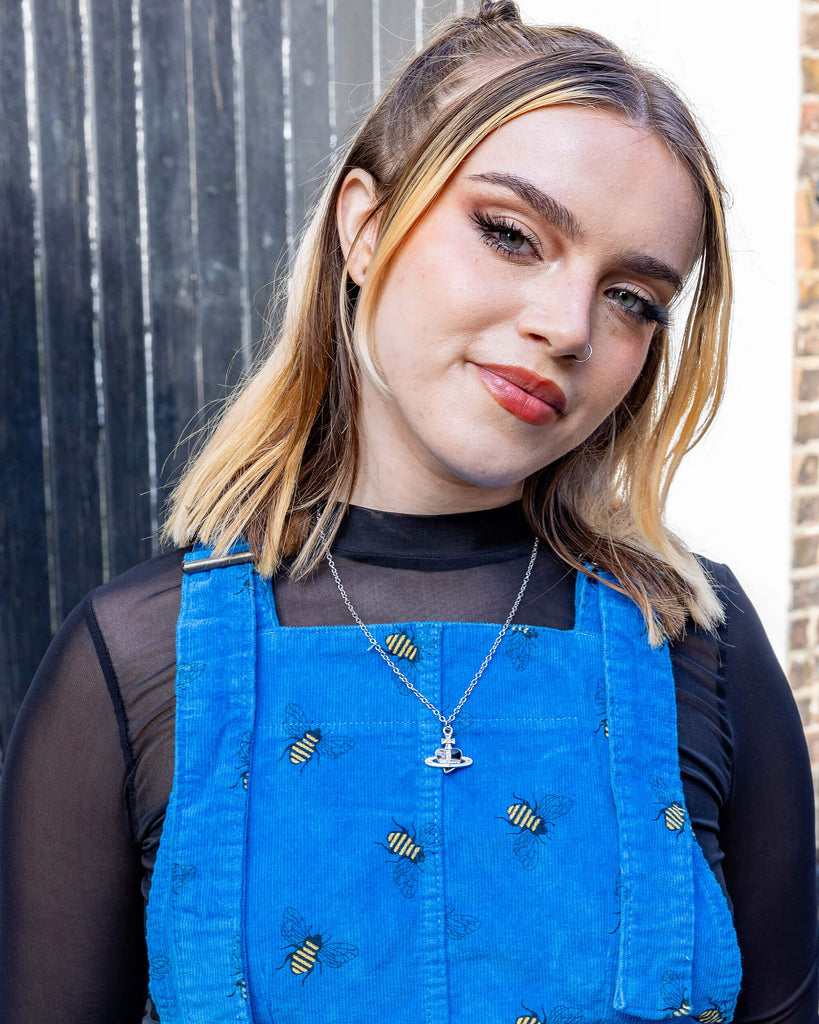 white femme model with blonde hair is wearing Run & Fly Blue Bee Stretch Corduroy Pinafore Dress paired with a long sleeve black mesh top. The bright blue pinafore has an all over bee print. The model is smiling at the camera with her hands behind her back outdoors in Hove.