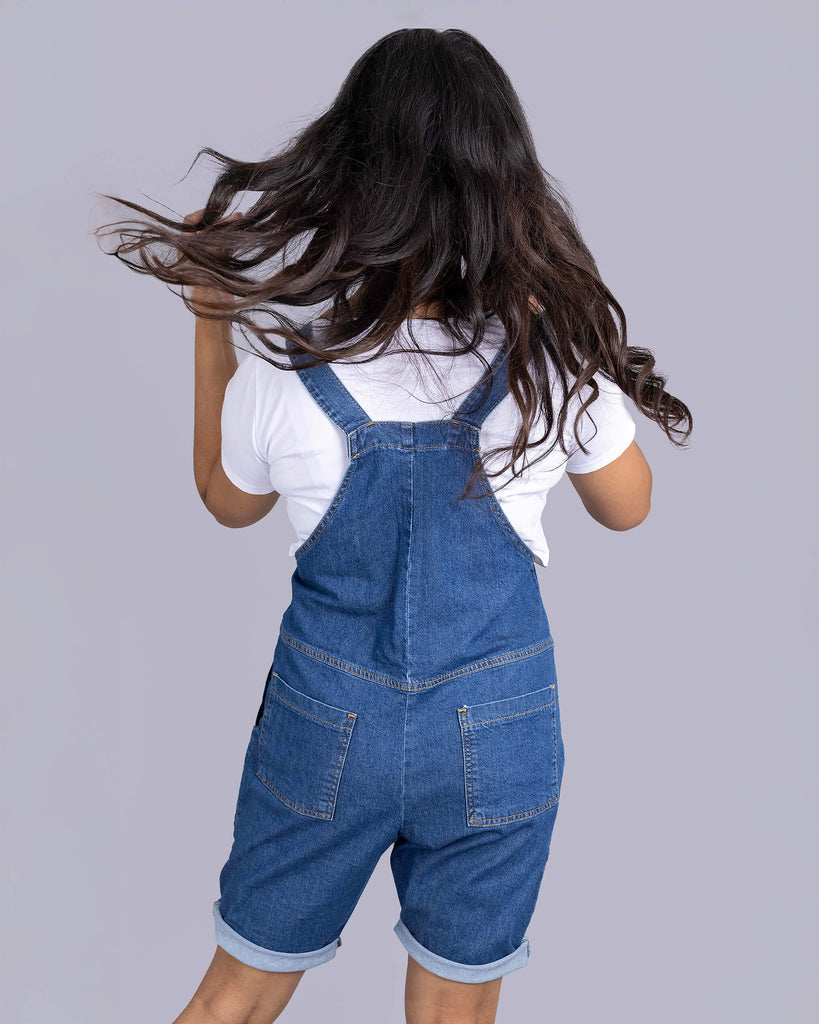 Beautiful Jarin in blue dungaree shorts and white tee underneath with white trainers. She is facing away from the camera flicking her hair up and off her shoulder to show off the back of the product.