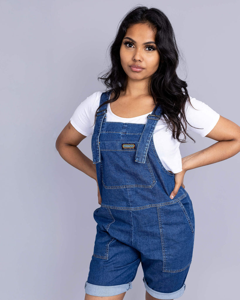 Beautiful Jarin in blue dungaree shorts and white tee underneath with white trainers. She has her hands in her pockets looking dead into camera and smiling.