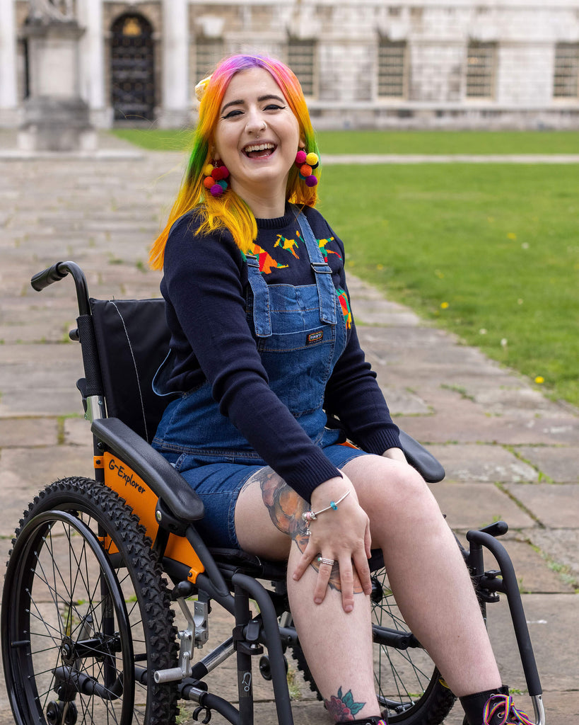 Rainbow haired femme person sat in a wheelchair  smiling into camera wearing blue stretch denim dungaree shorts and a blue rainbow dinosaur jumper underneath. 