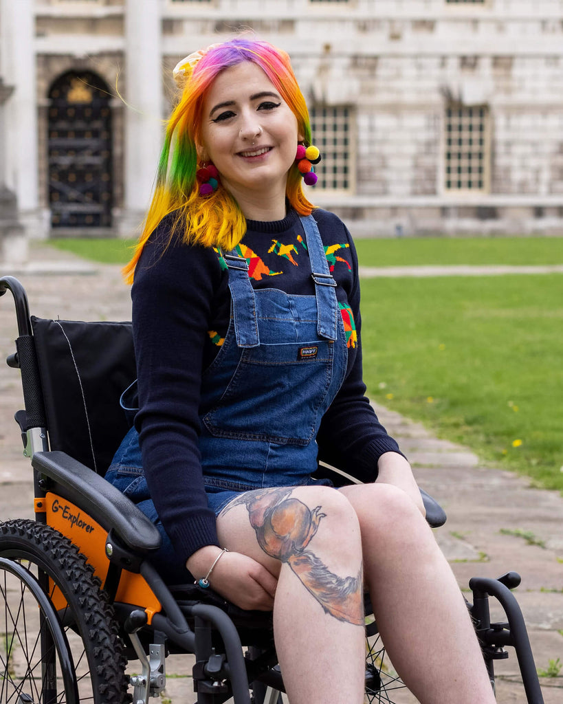 Rainbow haired femme person sat in a wheelchair smiling into camera wearing blue stretch denim dungaree shorts and a blue rainbow dinosaur jumper underneath.