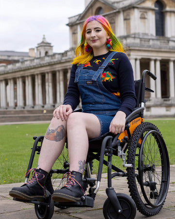 Rainbow haired femme person sat in a wheelchair smiling into camera wearing blue stretch denim dungaree shorts and a blue rainbow dinosaur jumper underneath.