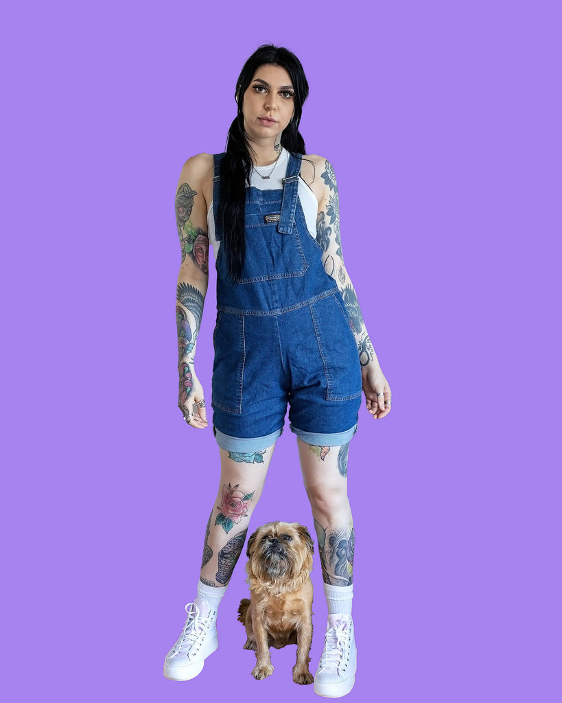 Rosie is stood on a pastel purple background. She is stood facing the camera with her hands resting by her side, she's wearing the Blue Stone Wash Denim Stretch Dungaree Shorts with a white vest top underneath and white socks with white trainers. There is also a small brown dog looking at the camera sat between Rosie's feet.