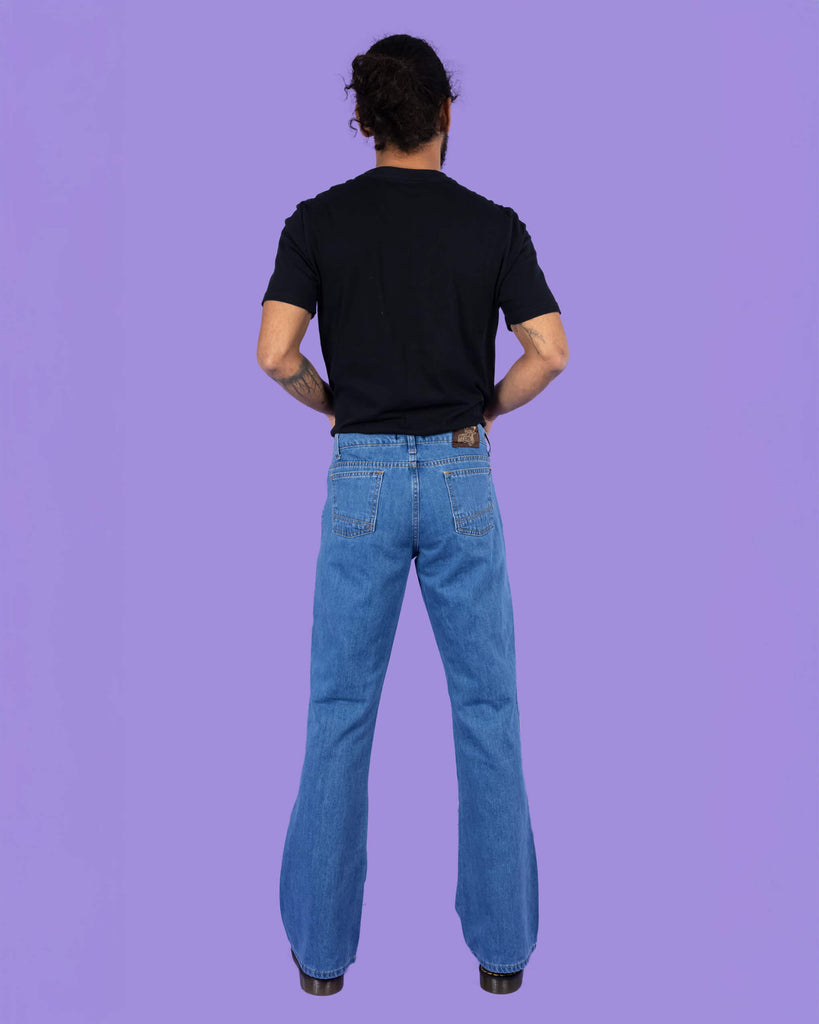 Richard, a hispanic male model with dark hair in a bun and a beard, is stood in a photography studio in Hove in front of a lilac backdrop wearing Stonewash Retro Boot Cut Flare Jeans with a black t shirt and black shoes. Richard is stood with his back to the camera and his arms in front of him.