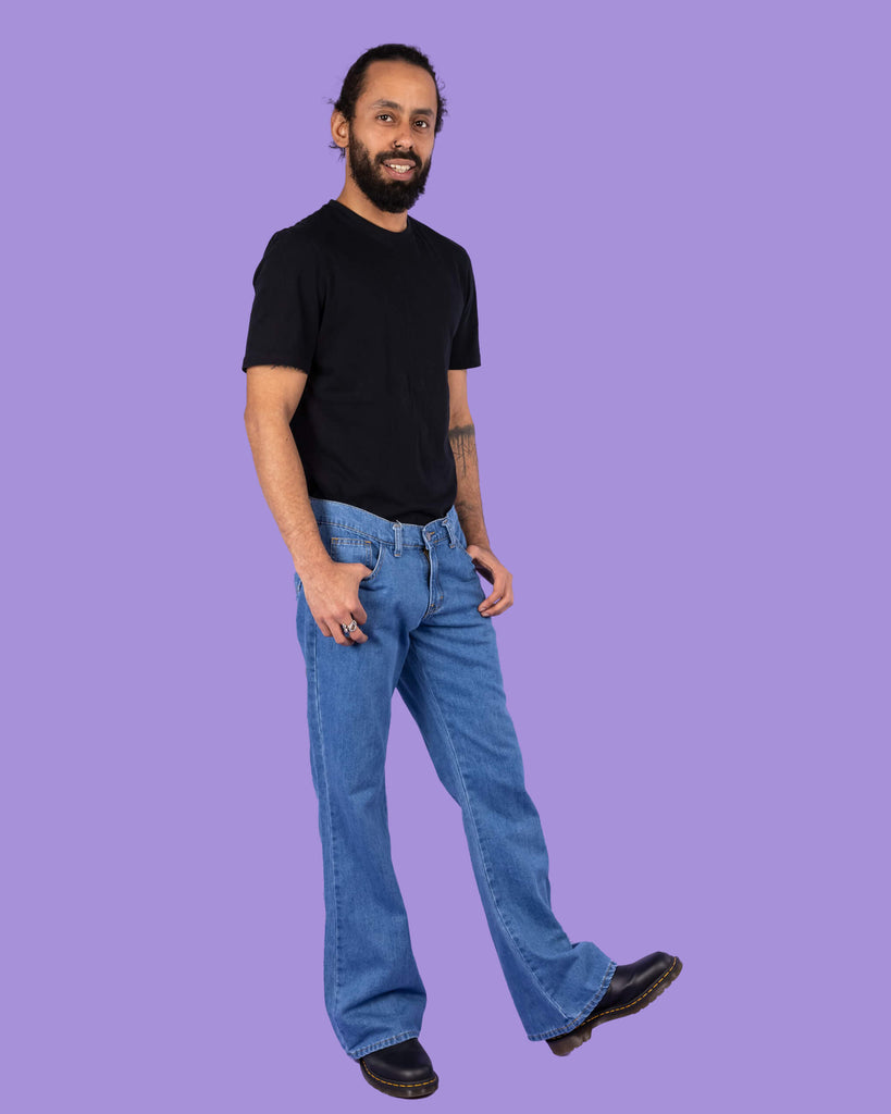 Richard, a hispanic male model with dark hair in a bun and a beard, is stood in a photography studio in Hove in front of a lilac backdrop wearing Stonewash Retro Boot Cut Flare Jeans with a black t shirt and black shoes. Richard is stood slightly to the side and is smiling with his thumbs resting in the front pockets of the jeans and one foot stretched outwards in front of him.