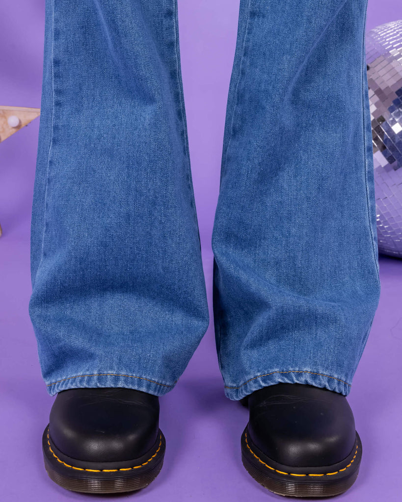 Close up of Richard wearing the Stonewash Retro Boot Cut Flare Jeans with black shoes. The background of the photo is lilac and there are disco balls behind him. The photo is cropped at the knees.