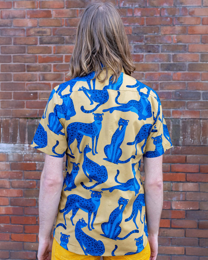 Jack is stood in front of a brick wall wearing the blue leopard short sleeve shirt with gold corduroy flares. They are facing away from the camera posing with both arms resting by their sides. Photo is cropped from the hips up.