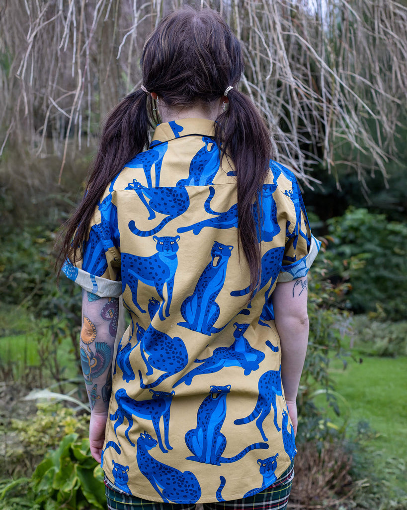 Florence is stood in front of a brick wall wearing the blue leopard short sleeve shirt with green tartan flares. They are facing forward posing with back to the camera. Photo is cropped from the hips up.