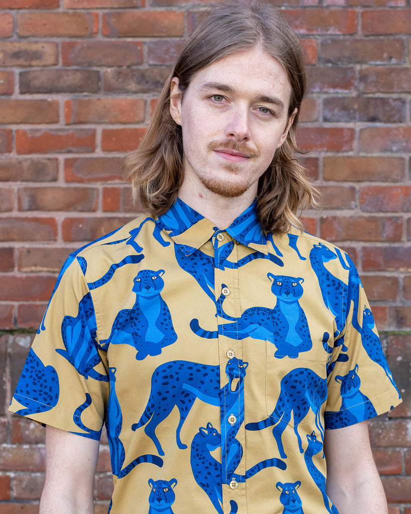 Jack is stood in front of a brick wall wearing the blue leopard short sleeve shirt with gold corduroy flares. They are facing forward posing with both hands resting by their sides and smiling whilst looking to camera. Photo is cropped from the waist up.