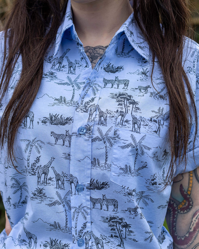 Flo is a femme dark haired model with tattoos she  is stood in front of a garden wearing the blue safari short sleeve shirt with tartan flares.
