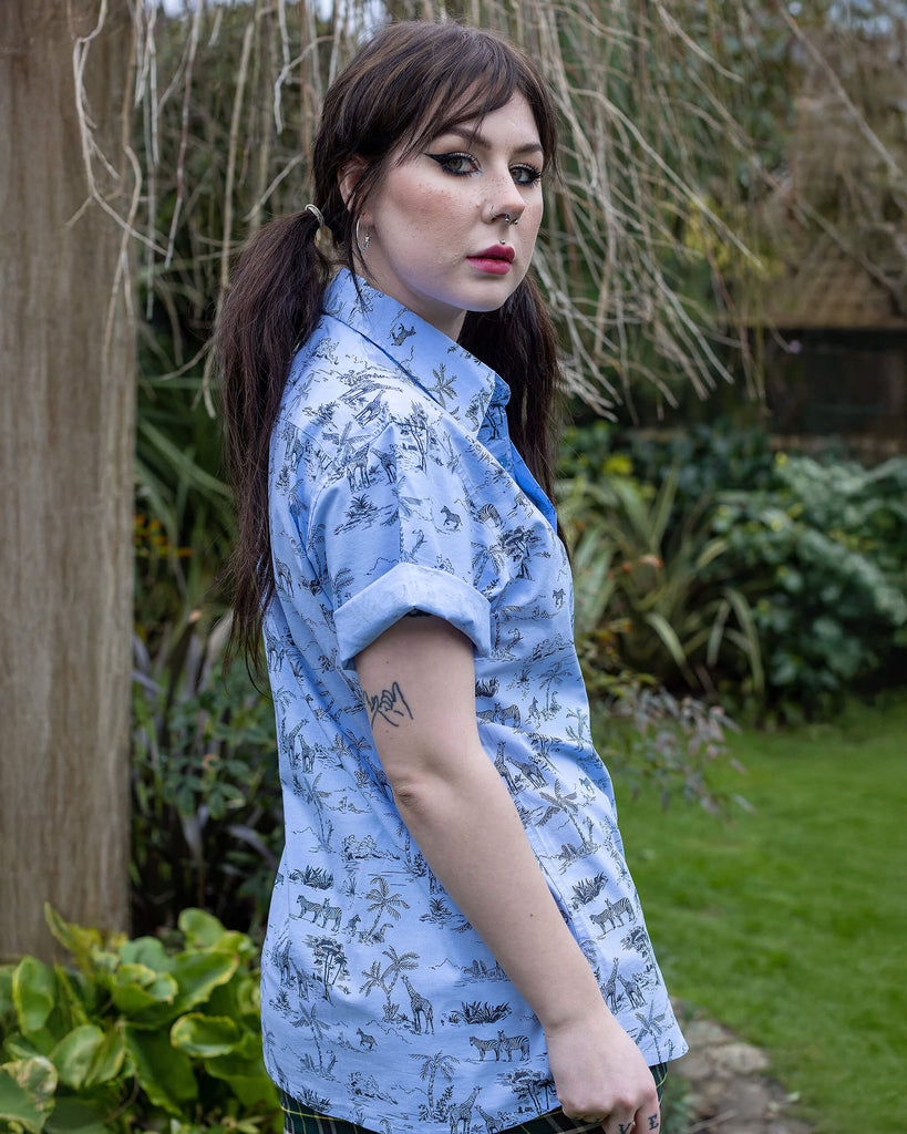 Flo is a femme dark haired model with tattoos she  is stood in front of a garden wearing the blue safari short sleeve shirt with tartan flares.