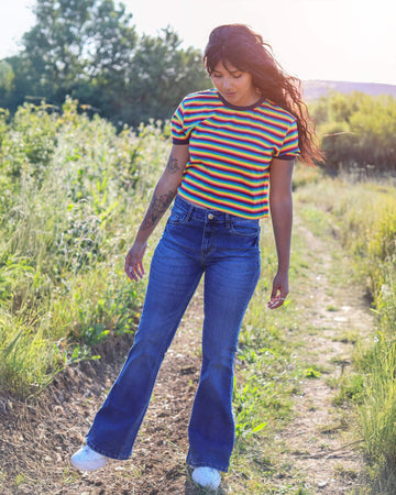 Meera is stood in a grassy field wearing the stone wash blue distress stretch denim rock n roll bell bottom flares with a retro rainbow short sleeve tshirt and white trainers. She is facing forward posing on one leg and both arms out to the sides whilst looking down.