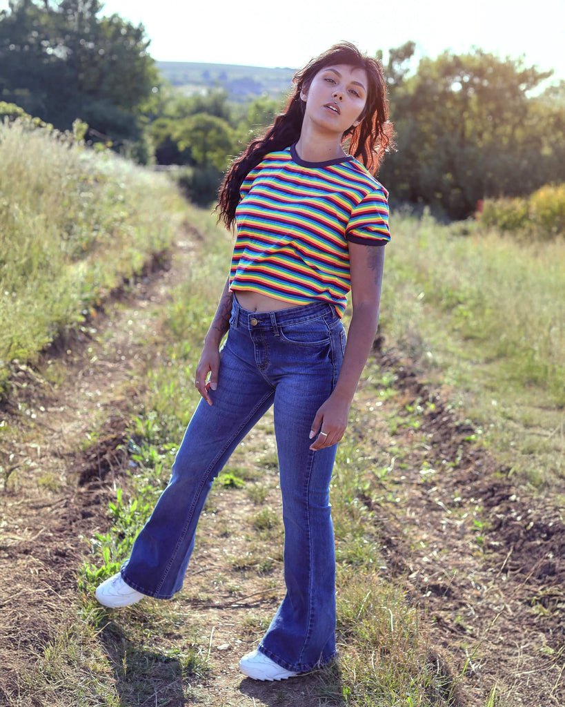 Meera is stood in a grassy field wearing the stone wash blue distress stretch denim rock n roll bell bottom flares with a retro rainbow short sleeve tshirt and white trainers. She is facing forward posing on one leg and leaning slightly back whilst looking to camera.